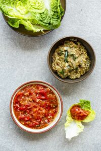 Rezeptfoto: Nam Prik Chili Relish mit Melanzani und Tomate in separaten Schalen, Salatblätter extra. Alles auf hellgrauem Untergrund.