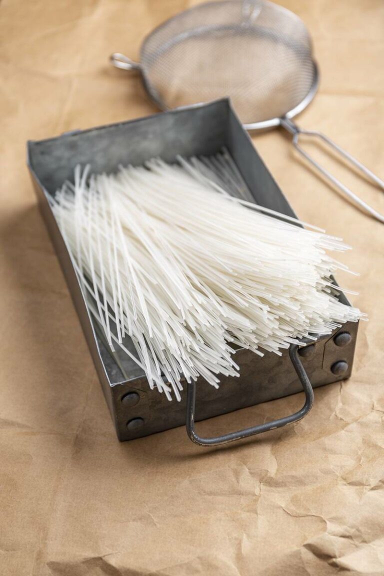 Moodfoto: Getrocknete Vermicelli Reisnudeln in eckigem Metallgeschirr. Im Hintergund ein Sieb, fotografiert auf Papieruntergrund.
